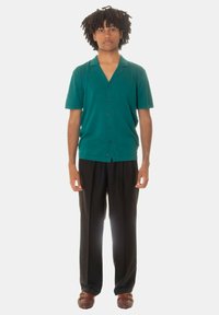Camicia corta a maniche corte di colore teal con colletto, chiusura a bottoni, abbinata a pantaloni neri e scarpe brown intrecciate. Design minimalista.