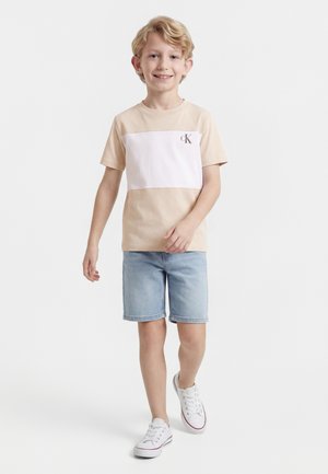 Usmívající se chlapec, který chodí, má na sobě béžovo-bílé tričko CK, světlé džínové kraťasy a bílé tenisky na jednobarevném bílém pozadí.
