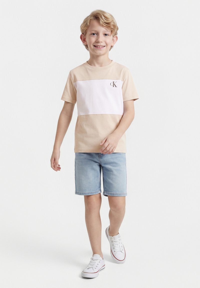 Χαμογελαστό αγόρι που περπατά, φοράει μπεζ και λευκό t-shirt CK, ανοιχτόχρωμα τζιν σορτς και λευκά αθλητικά παπούτσια σε απλό λευκό φόντο.