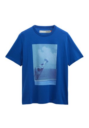 Μπλε κοντομάνικο μπλουζάκι με ξεθωριασμένη φωτογραφική εκτύπωση ενός χεριού που κρατά ένα μικρό κοχύλι με φόντο τον ουρανό και τη θάλασσα.