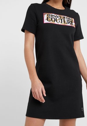 Vrouw met een korte zwarte jurk met het "Versace Jeans Couture"-logo in een bloemenlijst op de borst, staand met ontspannen armen langs de zijkanten.