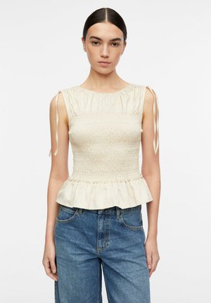 Donna che indossa un top crema senza maniche con arricciature, spalline con laccetti e orlo a peplo, abbinato a jeans di denim blu, su sfondo bianco.