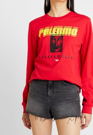 Osoba nosząca czerwoną koszulkę z długim rękawem z żółtym napisem „PALERMO”, czarną grafiką twarzy i czarnym napisem „LA CASA DE PAPEL”, zestawioną z czarnymi szortami dżinsowymi.