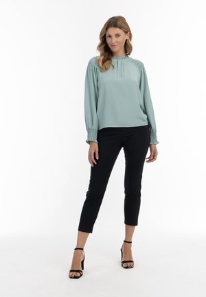 Bluză verde deschis cu decolteu plisat și manșete elasticizate, combinată cu pantaloni negri ajustați și tocuri negre cu curele. Textură de țesătură netedă.
