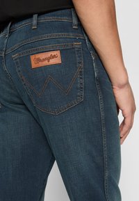 Tummansiniset farkut, joissa on nahkainen Wrangler-logo-etiketti takataskussa, erottuvalla keltaisella tikkaamalla ja istuvalla leikkauksella.