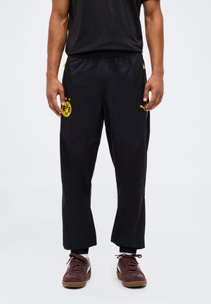 Man wearing black athletic pants with yellow BVB logo on left thigh and brown sneakers with white socks, standing against white background.