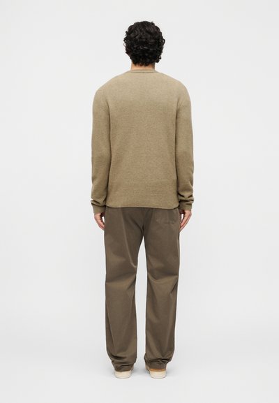 Άνδρας με σγουρά σκούρα μαλλιά, εικονιζόμενος από πίσω, φοράει μπεζ πουλόβερ, φαρδιά καφέ παντελόνια και ανοιχτόχρωμα παπούτσια επί απλοϊκού φόντου.