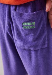 Poche d'un pantalon en éponge violet avec une broderie verte "American Vintage" et une partie d'une main reposant près de la ceinture.