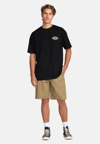 Sort bomulds T-shirt med et lille logo på brystet, parret med beige shorts og sorte sneakers med hvide detaljer.