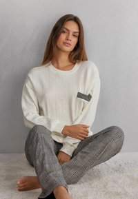 Hvid, ribbet langærmet top med en sort ternet lomme, parret med grå ternede pyjamasbukser, siddende på en lys tekstureret overflade.