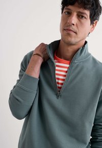 Grau zip-up Sweatshirt mit gerippten Bündchen, schichtweise über einem rot-weiß gestreiften Hemd getragen. Weicher Stoff, lässiges Design und ein Viertel-Längen-Reißverschluss.