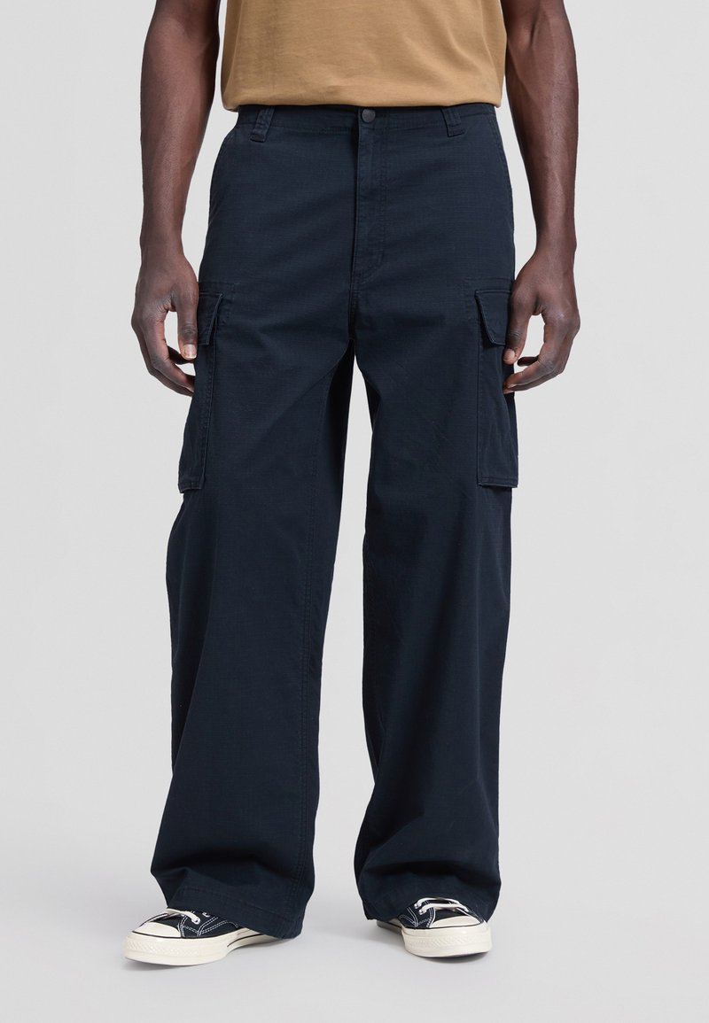 Homme portant un pantalon cargo large et sombre avec des poches à rabat et des baskets noires et blanches, debout devant un fond uni.
