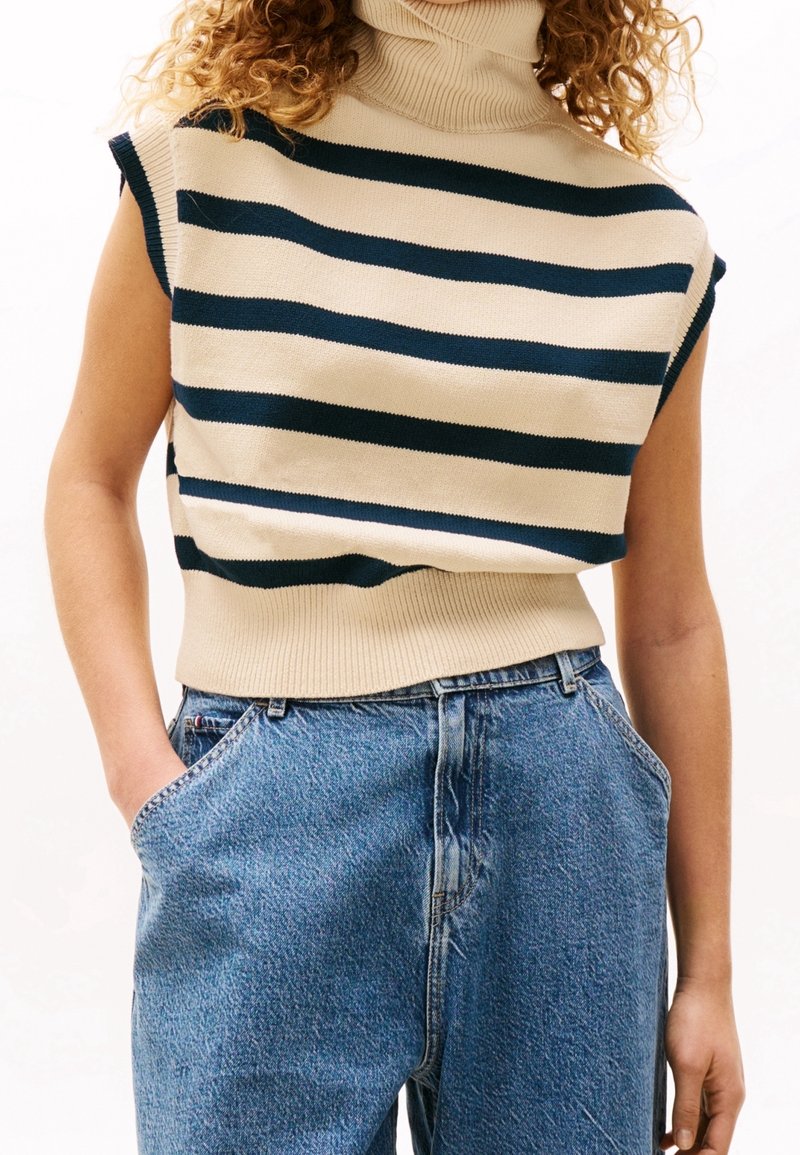 Sweter dziany z wysokim kołnierzem, w kremowo-granatowe poziome paski, z krótkimi rękawami i żebrowanym pasem; zestawiony z niebieskimi dżinsami.