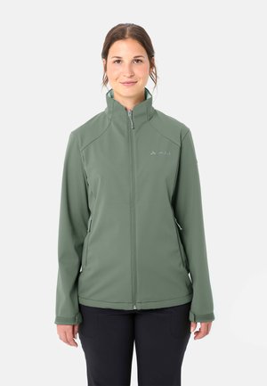 Femme portant une veste softshell olive à fermeture éclair, col montant et poches latérales zippées, debout devant un fond blanc.