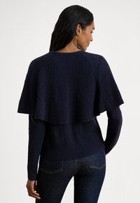 Tumesinine kootud kampsun kihilise seljatoega, sooniline tekstuur, pikad varrukad ja kroogitud alumine äär, kombineeritud tumedate teksadega.