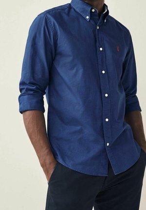 Mann trägt ein langärmliges, navyblaues Hemd mit Knöpfen, die Ärmel hochgekrempelt, und dunkle Hosen, die Hände in den Taschen.