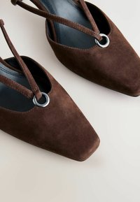 Zapatos de ante marrón con punta cuadrada, que cuentan con correas delgadas y un adorno metálico circular. Acabado interior liso, textura mínima.