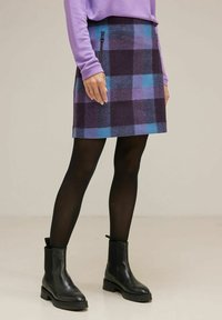 Kvinna klädd i en lila tröja och en rutiga kjol med blå och lila nyanser, kombinerat med svarta strumpbyxor och ankelstövlar, stående inomhus.