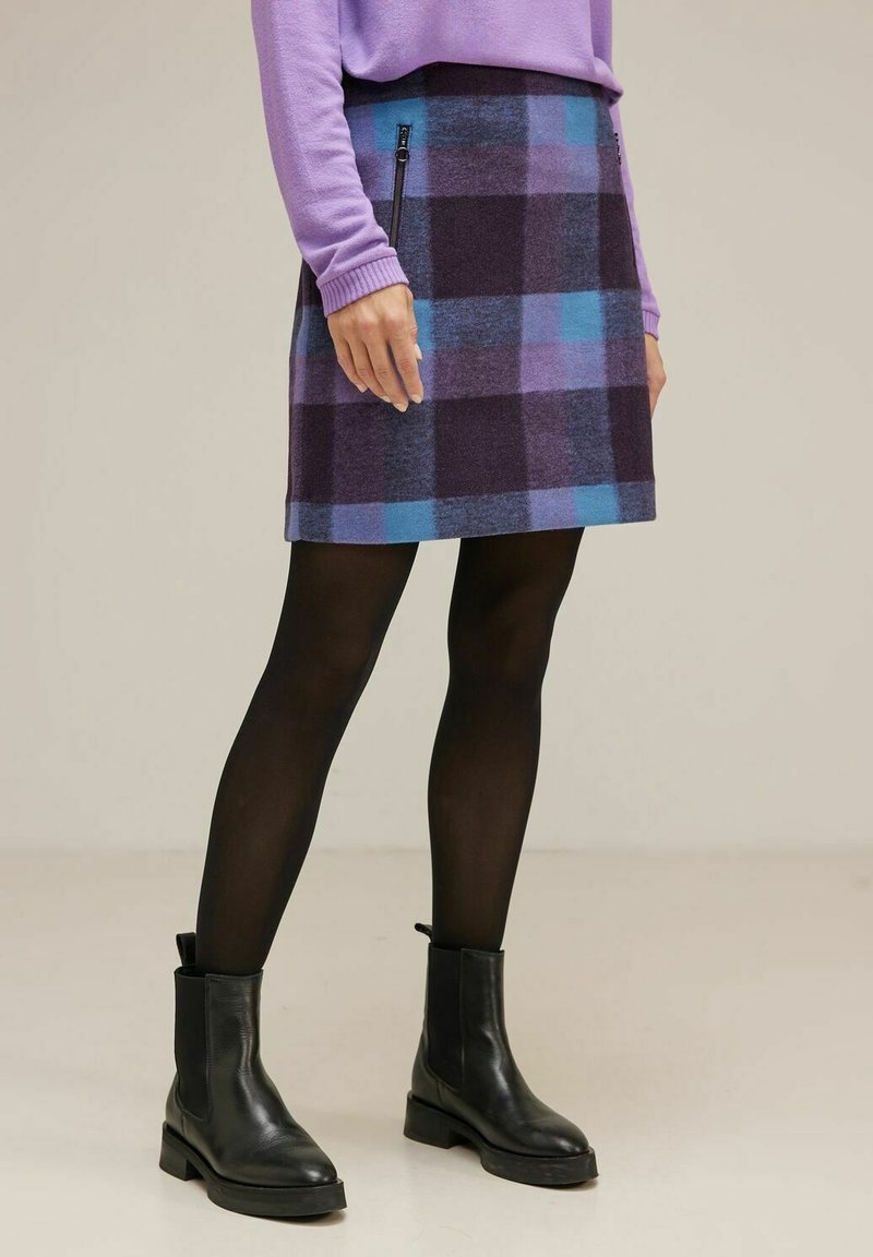 Kvinna klädd i en lila tröja och en rutiga kjol med blå och lila nyanser, kombinerat med svarta strumpbyxor och ankelstövlar, stående inomhus.