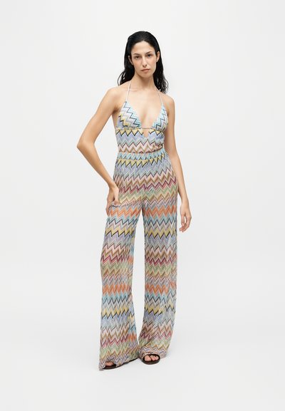 Femme debout dans une combinaison à motif zigzag coloré avec col halter, pantalon à jambes larges et sandales noires sur un fond blanc uni.