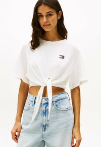 Camiseta blanca recortada con un detalle de nudo en la parte delantera, que presenta un pequeño logo impreso en el lado izquierdo del pecho. Llevada con jeans de tiro alto de color azul claro.