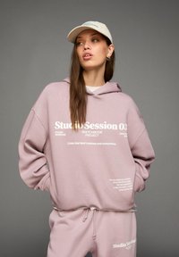 Vrouw in een lichtpaarse hoodie en bijpassende trainingsbroek met witte tekst, beige pet, staand met handen in de zakken tegen een grijze achtergrond.