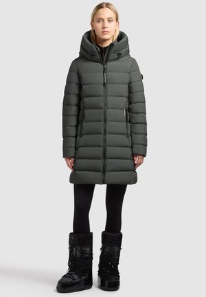 Donkergroen puffercoat met een hoge kraag en grote capuchon, voorzien van een rits aan de voorkant en zijzakken. Zwarte waterdichte laarzen met veters.