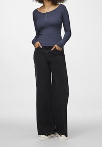 Frau mit lockigem Haar, die ein marineblaues Langarm-Henley-Oberteil, weite schwarze Jeans und weiße Plateausneaker trägt und mit den Händen in den Taschen steht.