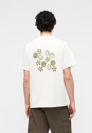 Mężczyzna ubrany w białą koszulkę z zielonymi i beżowymi wzorami parasoli na plecach, w brązowych krótkich spodenkach typu cargo, stoi tyłem.