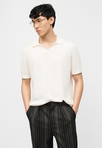 Νεαρός άνδρας με στρογγυλά γυαλιά, φόρεμα κρεμ κοντομάνικου πόλο και μαύρο παντελόνι με λεπτές κάθετες μπεζ ρίγες, με τα χέρια στις τσέπες.