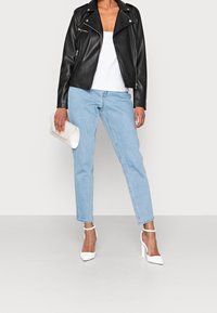 Chaqueta de cuero negra sobre una camiseta sin mangas blanca, combinada con jeans rectos de color azul claro y zapatos de tacón blancos. Sosteniendo un bolso de mano blanco.