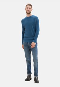 Suéter de punto azul con un cuello redondo, ajuste ceñido. Combinado con unos jeans de un azul claro y botas negras hasta el tobillo. Textura lisa, sin patrones visibles.