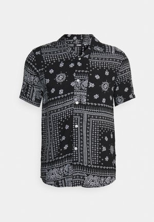 Homme portant une chemise à manches courtes avec un motif noir et blanc, un pantalon gris clair retroussé et des baskets blanches, debout, une main dans la poche.