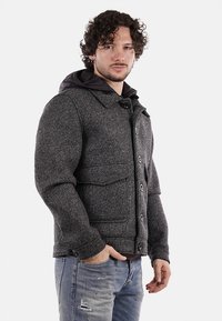 Giacca grigia texture con cappuccio, dotata di chiusura a bottoni, due tasche sul petto e polsini a costina. Indossata con jeans chiari strappati.
