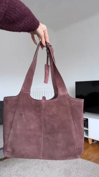 Hand håller en stor mauve mocka toteväska med två handtag i ett vardagsrumsmiljö.