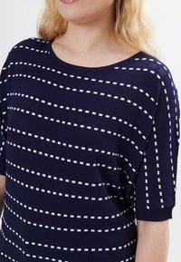 Maglione lavorato a maglia blu navy con righe orizzontali bianche e motivi a quadrati. Presenta un colletto rotondo e maniche corte. Texture liscia.