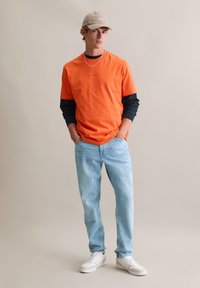 Pomarańczowy bawełniany t-shirt nałożony na ciemną bluzę z długim rękawem, do tego jasnoniebieskie jeansy i białe trampki. Całość dopełnia beżowa czapka z daszkiem.