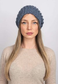Pletená modrá čepice s texturovaným žebrováním, s obroubeným okrajem. Nesešena osobou s dlouhými vlasy, v kombinaci s béžovým svetrem.
