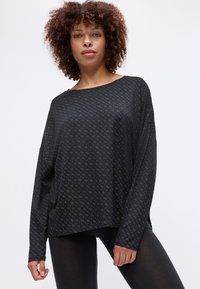 Mulher com cabelo encaracolado vestindo uma blusa preta de manga longa solta com um padrão geométrico sutil e leggings pretas, contra um fundo simples.