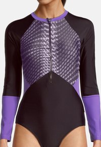 Maillot de bain à manches longues en noir et violet. Présente un panneau texturé violet avec un motif géométrique et une fermeture éclair centrale. Fini en tissu lisse.