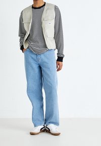 Beige verktygsväst över en svart och vit randig långärmad tröja, kombinerat med ljusblå vida jeans och vita sneakers.