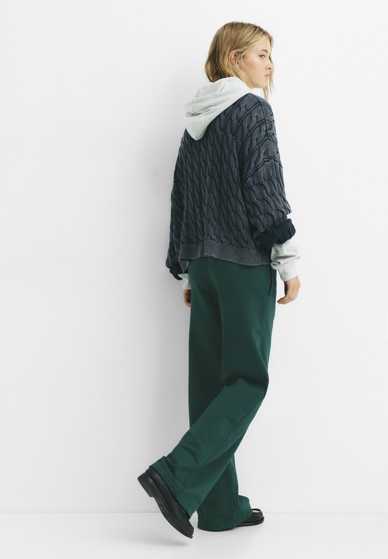 Chunky strikket sweater i navy med lysegrå hættetrøje underneath, parret med brede grønne bukser og sort fodtøj.