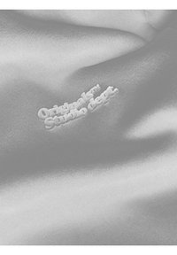 Ljusgrå satin som har ett präglat vitt logo med texten "Originals Studio dept." i ett lekfullt, böjt typsnitt.