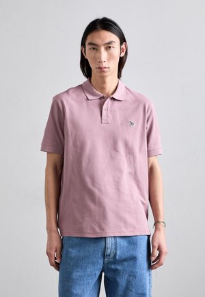 Camiseta polo de algodón en rosa claro con cuello, mangas cortas y un pequeño logotipo bordado en el pecho. Combinada con jeans de mezclilla azul.