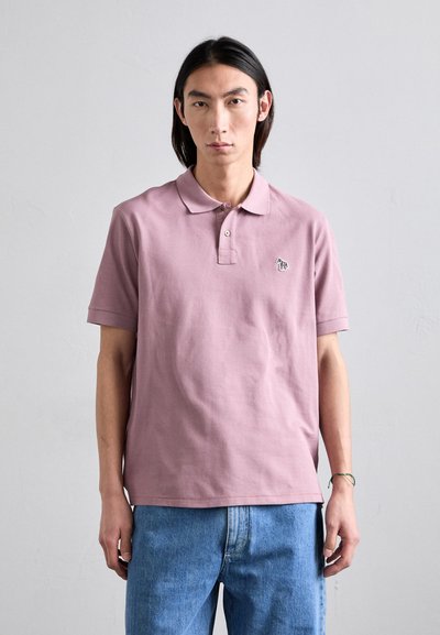 Camiseta polo de algodón en rosa claro con cuello, mangas cortas y un pequeño logotipo bordado en el pecho. Combinada con jeans de mezclilla azul.