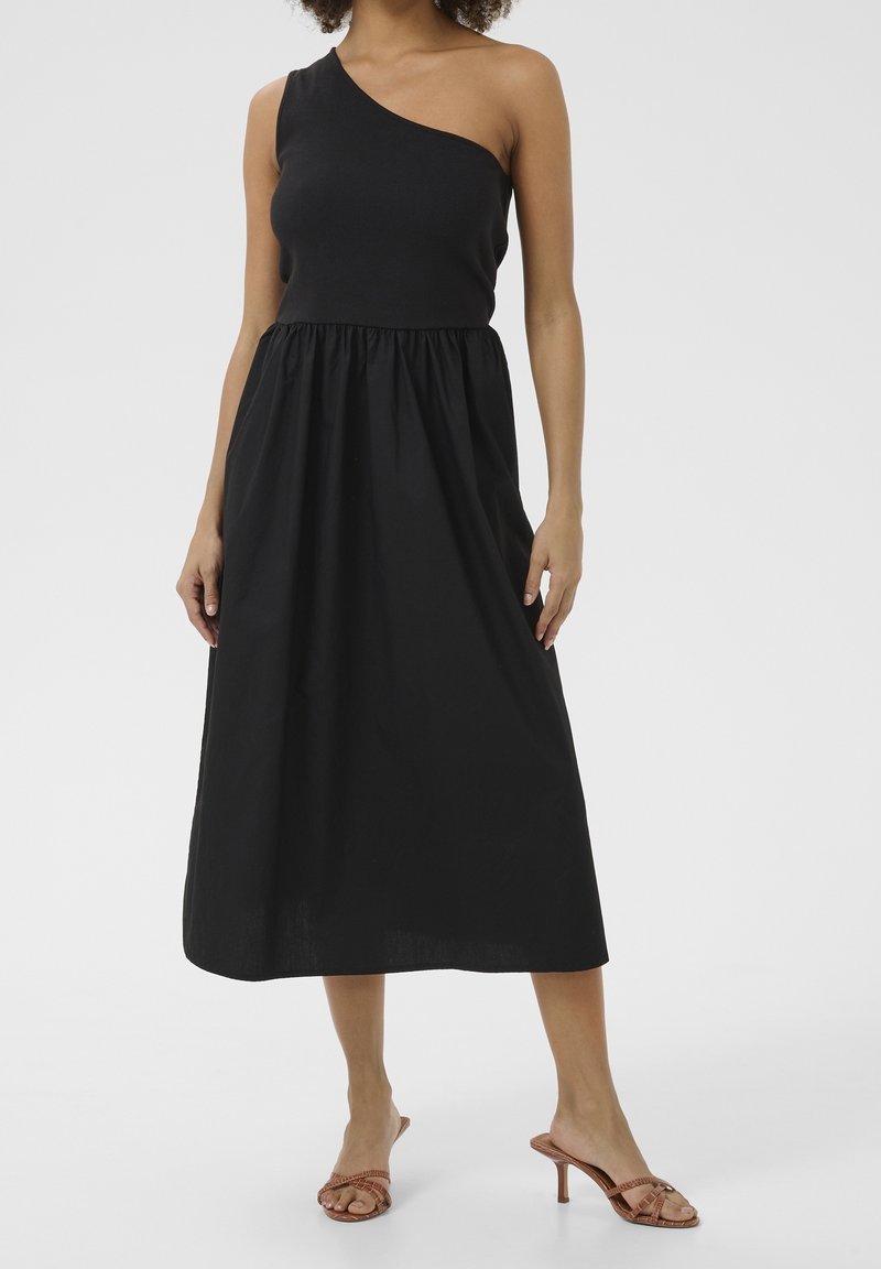Femme portant une robe midi noire asymétrique avec une taille froncée et des sandales à talons marron, debout devant un fond uni.
