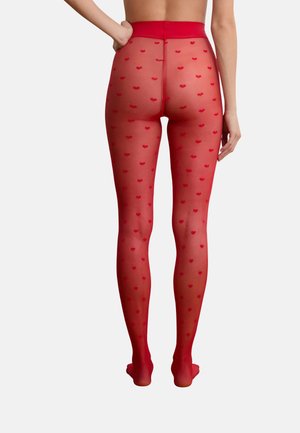 Femme portant des collants rouges transparents avec de petits motifs en forme de cœur, vue de dos sur un fond uni.