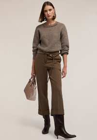 Kvinna i en brun stickad tröja, högmidjade vida byxor med ett rep-bälte, mörkbruna stövlar, som håller en beige vadderad handväska.