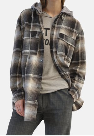 Personne portant un sweat à capuche gris sous une chemise à carreaux noire et marron boutonnée, assortie à un pantalon à carreaux gris foncé et une bague en argent à la main gauche.