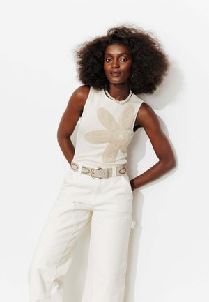 Femme aux cheveux bouclés et volumineux portant un haut blanc sans manches avec un grand motif floral, un pantalon blanc taille haute, une ceinture et un collier de perles.